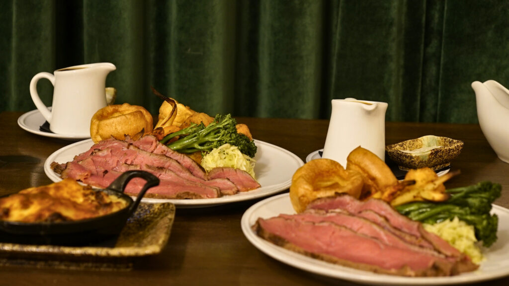 Hurley House - Three plates of roast beef with Yorkshire pudding, broccoli, and potatoes on a brown table, accompanied by two white gravy pitchers and a small dish of butter, set against a green velvet curtain background.