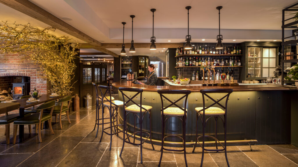 Hurley House - A cozy, warmly lit bar with high chairs around a curved wooden counter, shelves stocked with bottles, and a bartender behind the bar. There are tables and chairs to the left, with golden lights and rustic decor throughout.