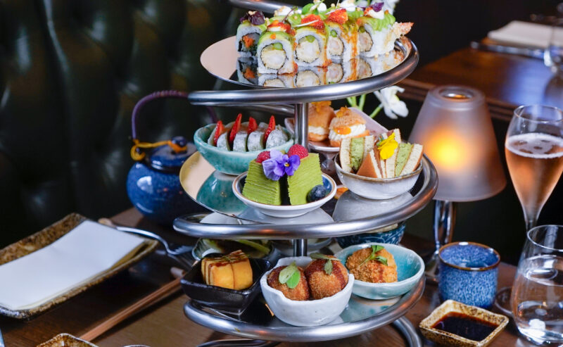 Hurley House - A three-tiered serving tray holds assorted sushi rolls, Japanese desserts, and small savory bites, surrounded by a teapot, glasses of champagne, and dining tableware in an elegant restaurant setting.