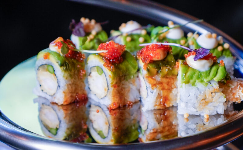 Hurley House - A close-up of colorful sushi rolls topped with avocado slices, red fish roe, sauce, and garnishes, arranged on a reflective silver plate.