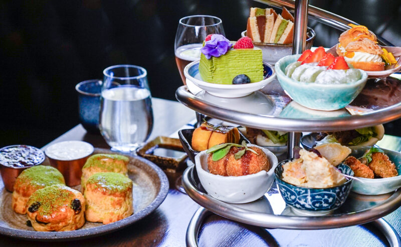 Hurley House - A tiered stand holds assorted desserts and snacks, including cakes, pastries, and fruit-topped treats. Scones with jam and cream, plus glasses of water, are set on the table, suggesting an afternoon tea setting.