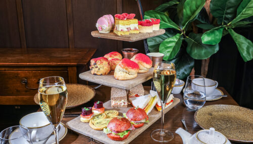 Hurley House - A three-tiered afternoon tea stand holds assorted sandwiches, scones, and colorful pastries. Two glasses of champagne, tea cups, and plates surround the stand on a wooden table, with green plants in the background.
