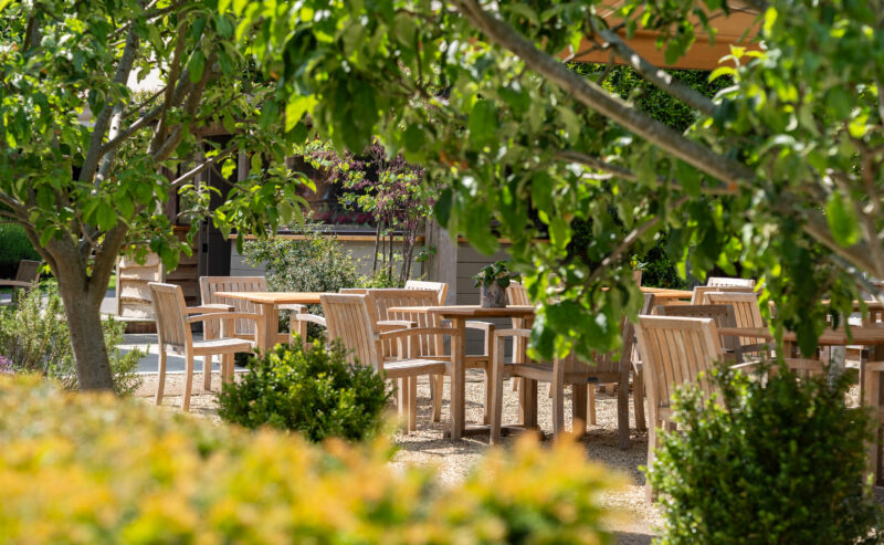 Hurley House - Wooden tables and chairs are arranged outdoors in a garden patio area, surrounded by green trees and shrubs on a sunny day.