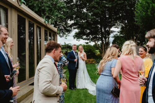 Hurley House - A bride and groom stand together smiling outside as wedding guests in colorful outfits gather around them, holding drinks and chatting on a grassy lawn near a building and trees.