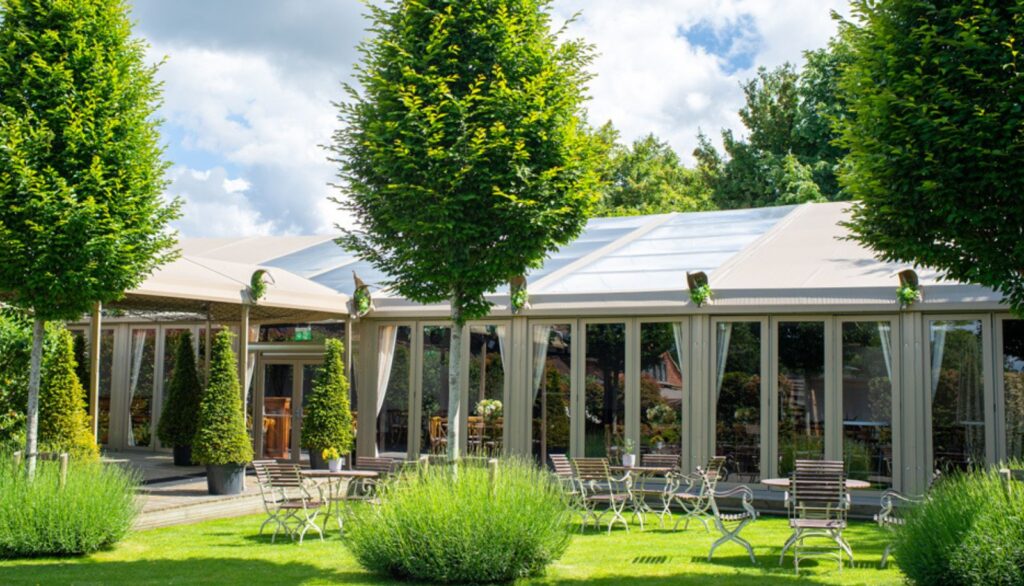 Hurley House - A luxury wedding venue in Berkshire features a charming outdoor dining area with tables and chairs set on lush green grass, surrounded by neatly trimmed bushes and tall trees. Large windows on the elegant building reflect the partly cloudy sky above.