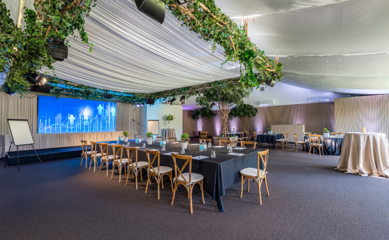 Hurley House - A modern conference room with U-shaped tables, wooden chairs, a large digital screen displaying graphs, greenery hanging from the ceiling, and cocktail tables in the background. The space is bright and elegant.