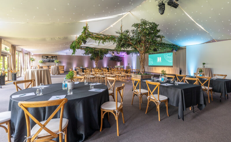 Hurley House - A spacious, elegant event hall with round and rectangular tables, black tablecloths, wooden chairs, indoor trees, and a ceiling decorated with lights and greenery. A presentation screen is visible at the front.