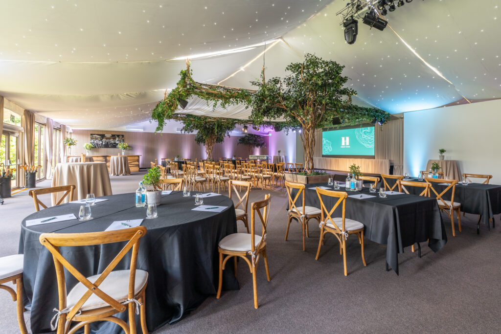 Hurley House - Spacious event hall at the premier meeting & conference venue in Berkshire, featuring round and rectangular tables with black cloths, wooden chairs, decorative greenery, soft lighting, a large screen, and ceilings draped in white fabric and fairy lights.