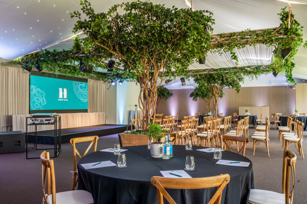 Hurley House - A spacious event room with round tables and wooden chairs, decorated with large indoor trees and greenery. A stage with a screen displaying an “H” logo is set up at the front. Glasses, water bottles, and notepads are on the tables.