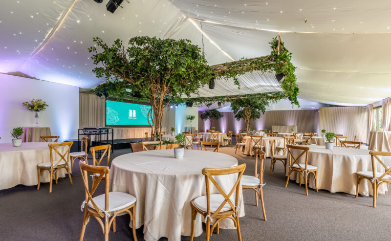 Hurley House - A spacious, elegant event room with round tables covered in beige cloths, wooden chairs, potted plants, and large indoor trees. Soft lighting and draped ceiling fabric with string lights create a cozy atmosphere. A video screen is at the front.