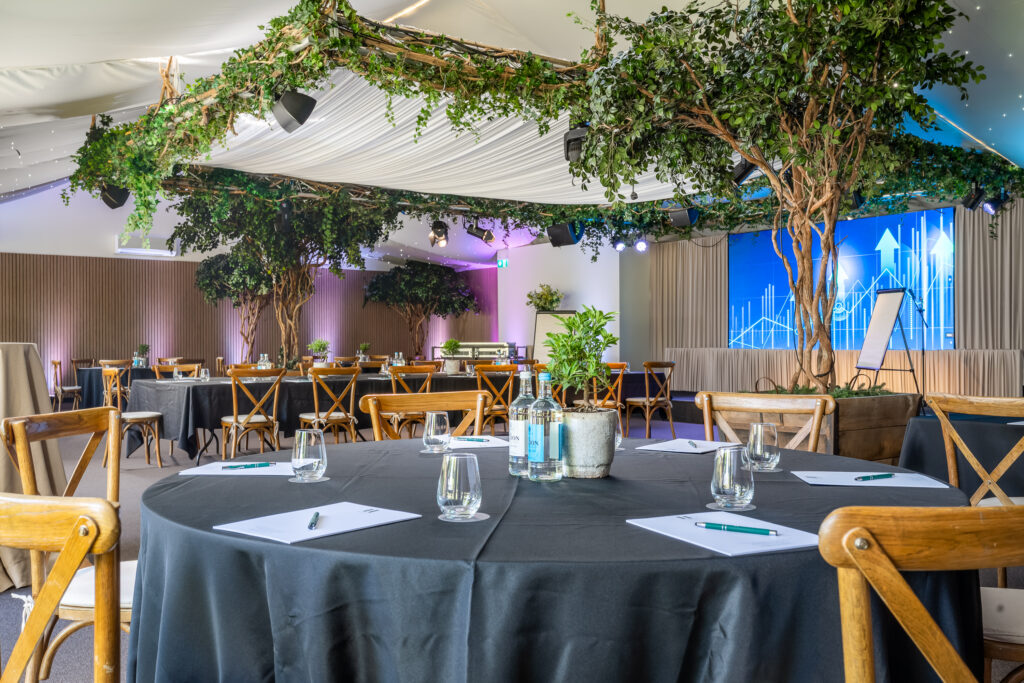 Hurley House - A conference room with round tables covered in black tablecloths, set with notepads, pens, glasses, and bottled water. Large artificial trees and greenery decorate the space, and a screen with a graph is visible in the background.