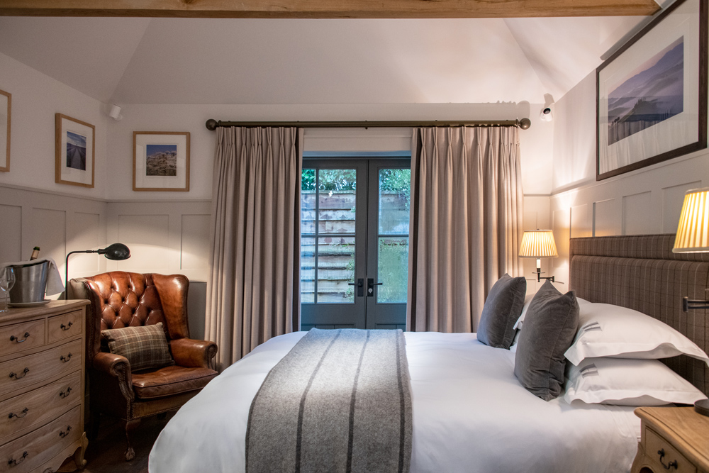 Hurley House - A cozy bedroom in a boutique wedding venue in Berkshire features a large bed with gray blankets and multiple pillows. A brown leather armchair sits beside a wooden dresser with a lamp. Framed pictures adorn the walls, and curtains cover glass doors leading outside.