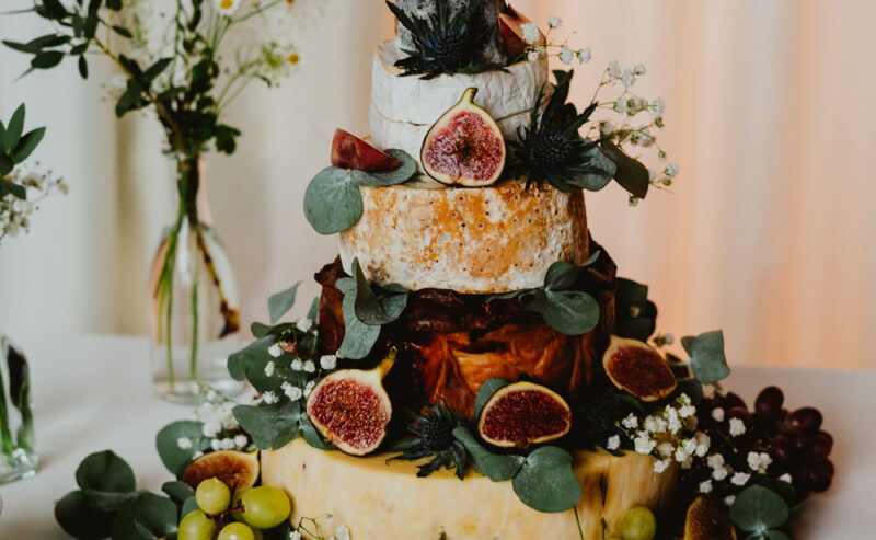 Hurley House - A three-tiered cheese cake is adorned with figs, grapes, and eucalyptus leaves, epitomizing the elegance of a luxury wedding venue in Berkshire. Encircled by small boxes of crackers on a white tablecloth, it sits alongside vases filled with greenery and white flowers.