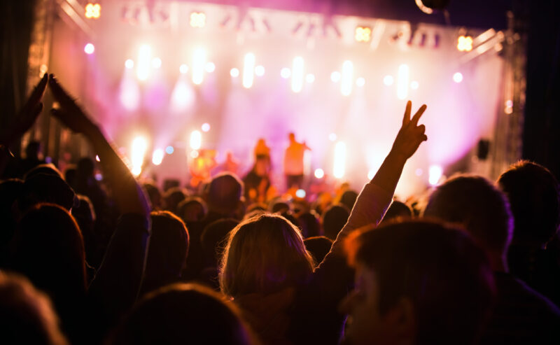 Hurley House - A crowd at a concert with colorful stage lights in the background; one person in the foreground is raising their hand and making a peace sign—capturing the excitement of whats on at Hurley House.