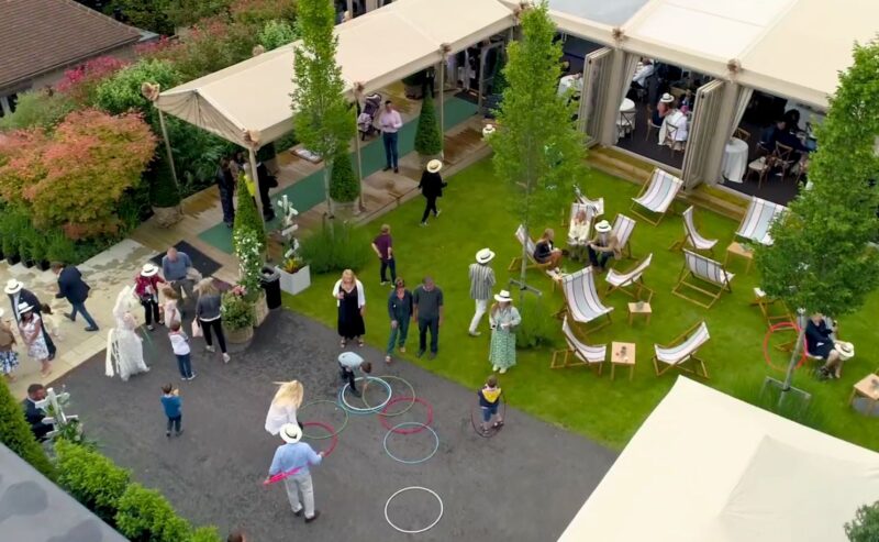 Hurley House - Aerial view of an outdoor garden party. People are socializing near tents and playing with hula hoops. Relaxing in deck chairs and mingling on the lawn. The setting is surrounded by greenery, with a casual, festive atmosphere.