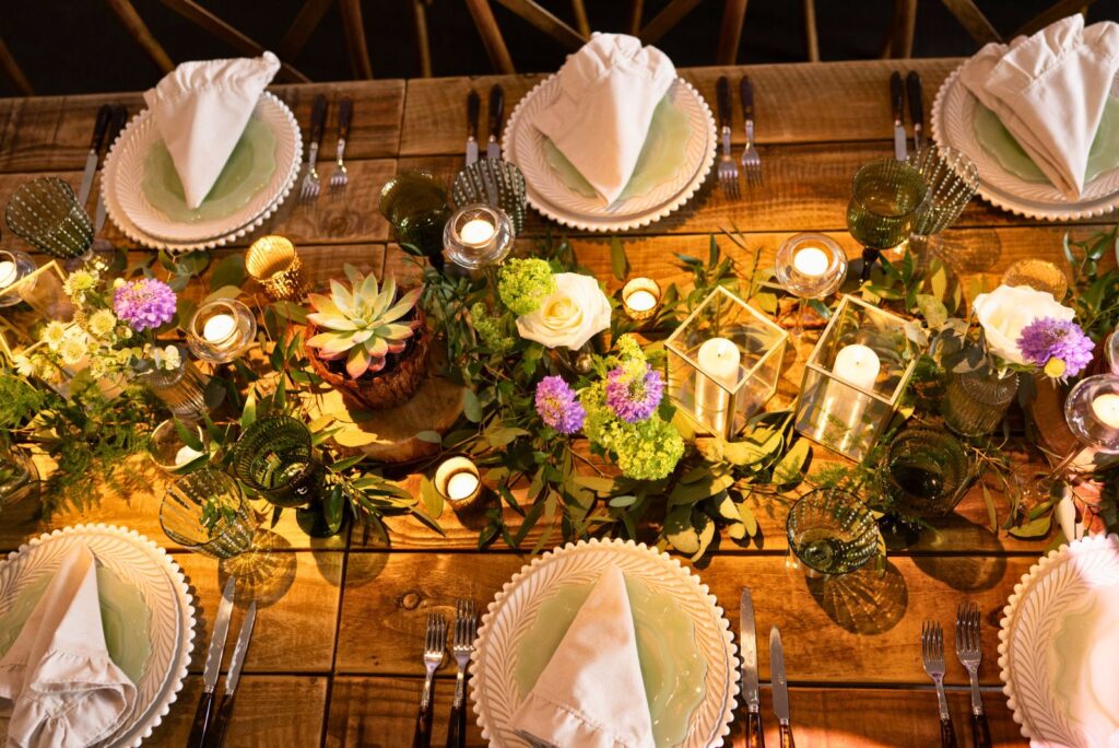 Hurley House - A rustic wooden table at this luxury wedding venue in Berkshire is elegantly set with white plates, neatly folded napkins, and green-tinted glasses. The centerpiece features candles, succulents, and a mix of purple and green flowers, creating a warm, inviting atmosphere.